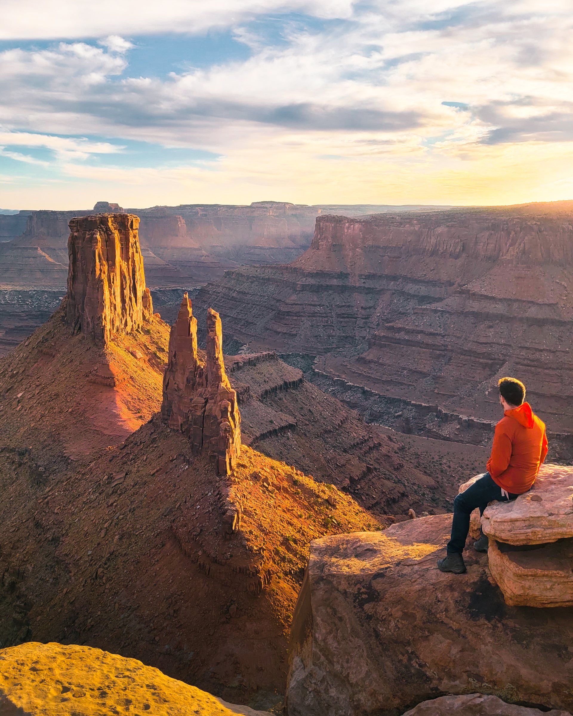 Hiker in a red jacket sitting on a sandstone ledge above a canyon of red rock towers at sunset