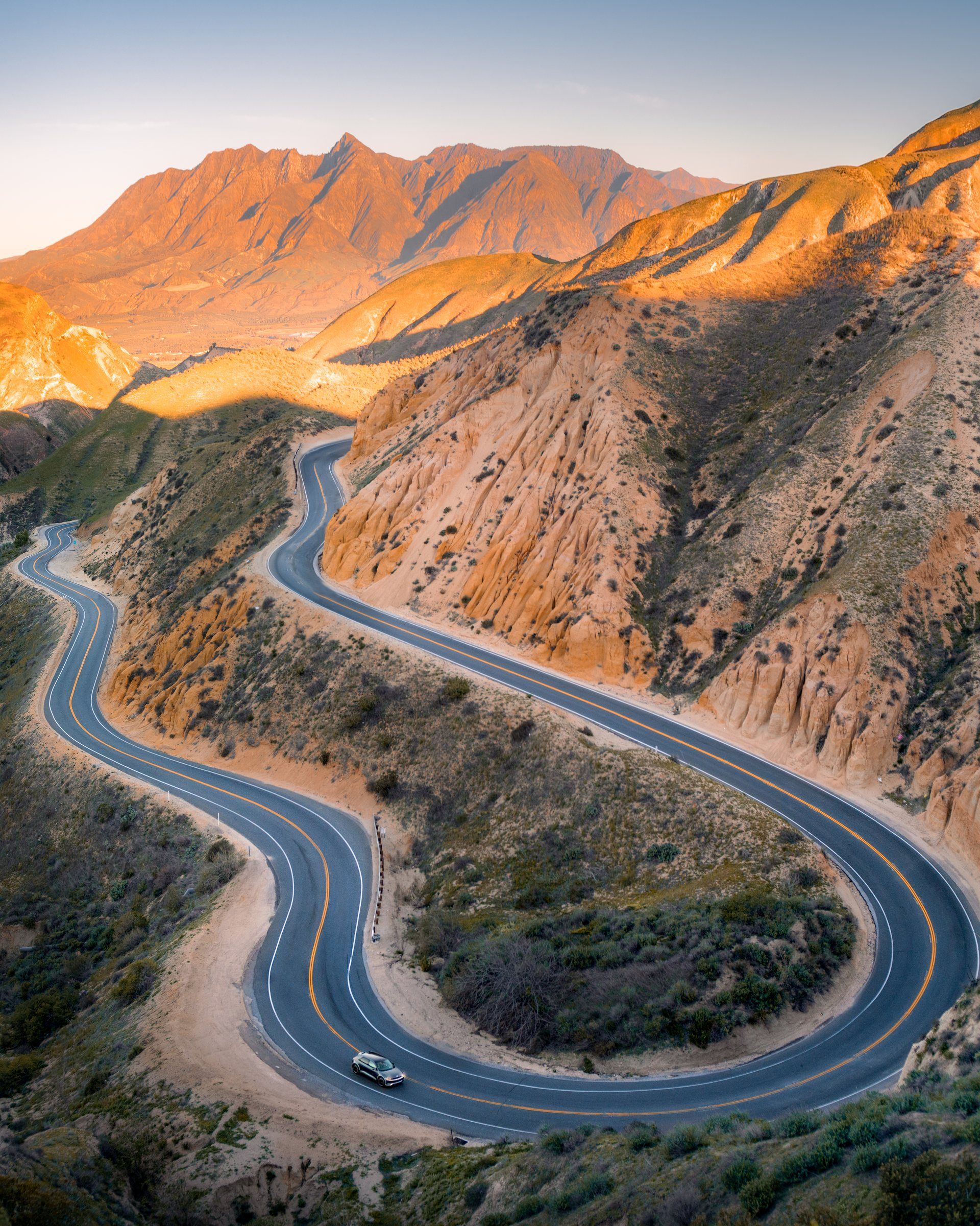 A Hyundai Ioniq 5 on a winding canyon road through sunlit badlands below rugged peaks