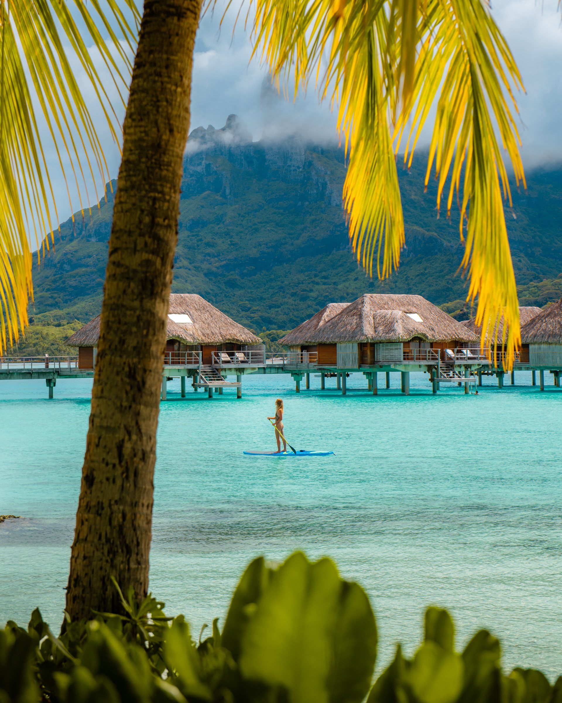 A paddleboarder on the turquoise lagoon in front of thatched overwater bungalows, framed by palms, with Mount Otemanu behind