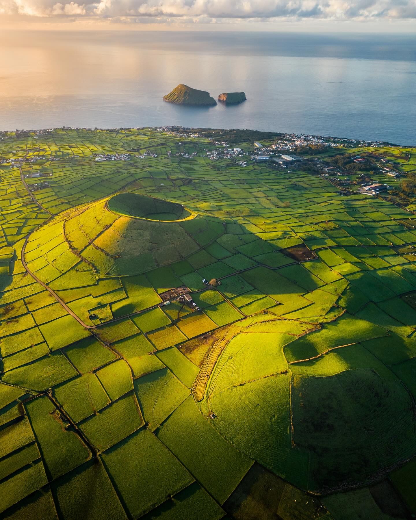 Aerial of a green volcanic cone in a patchwork of stone-walled fields, with two islets offshore at sunrise