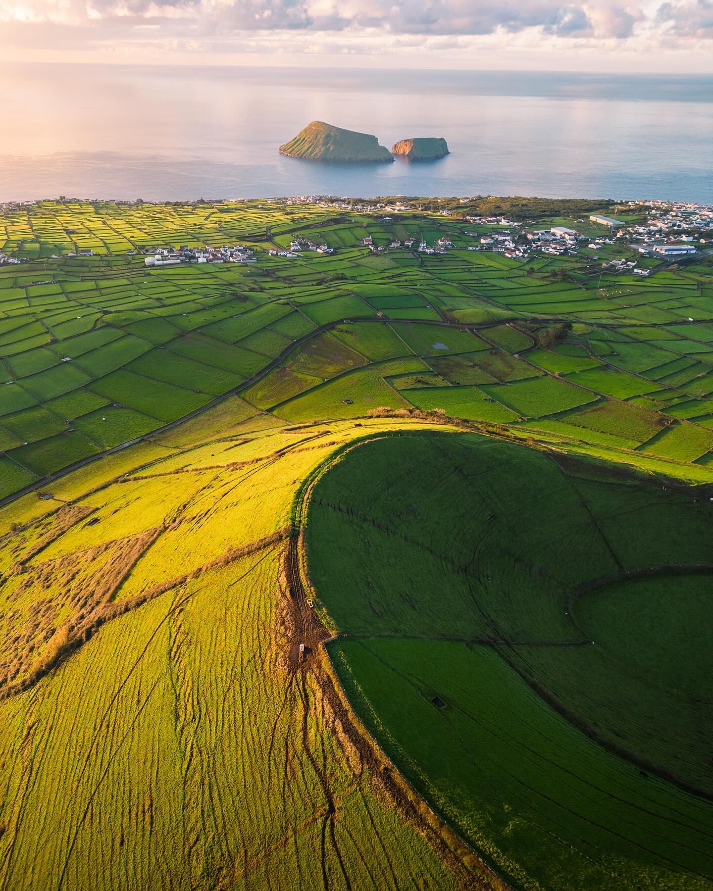 The rim of a grassy crater above patchwork fields and a coastal town, islets in the sea beyond