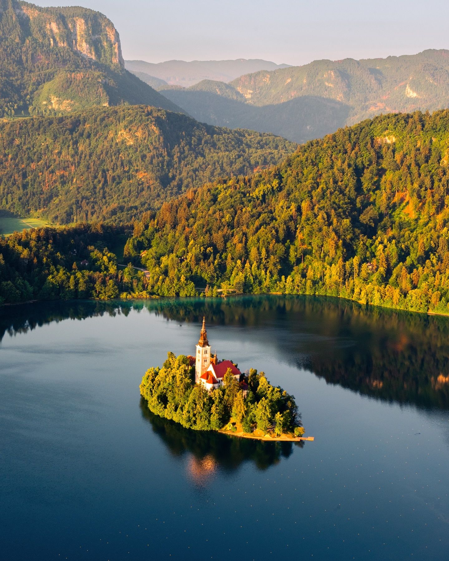 Aerial of the island church on Lake Bled in golden morning light, with forested hills behind