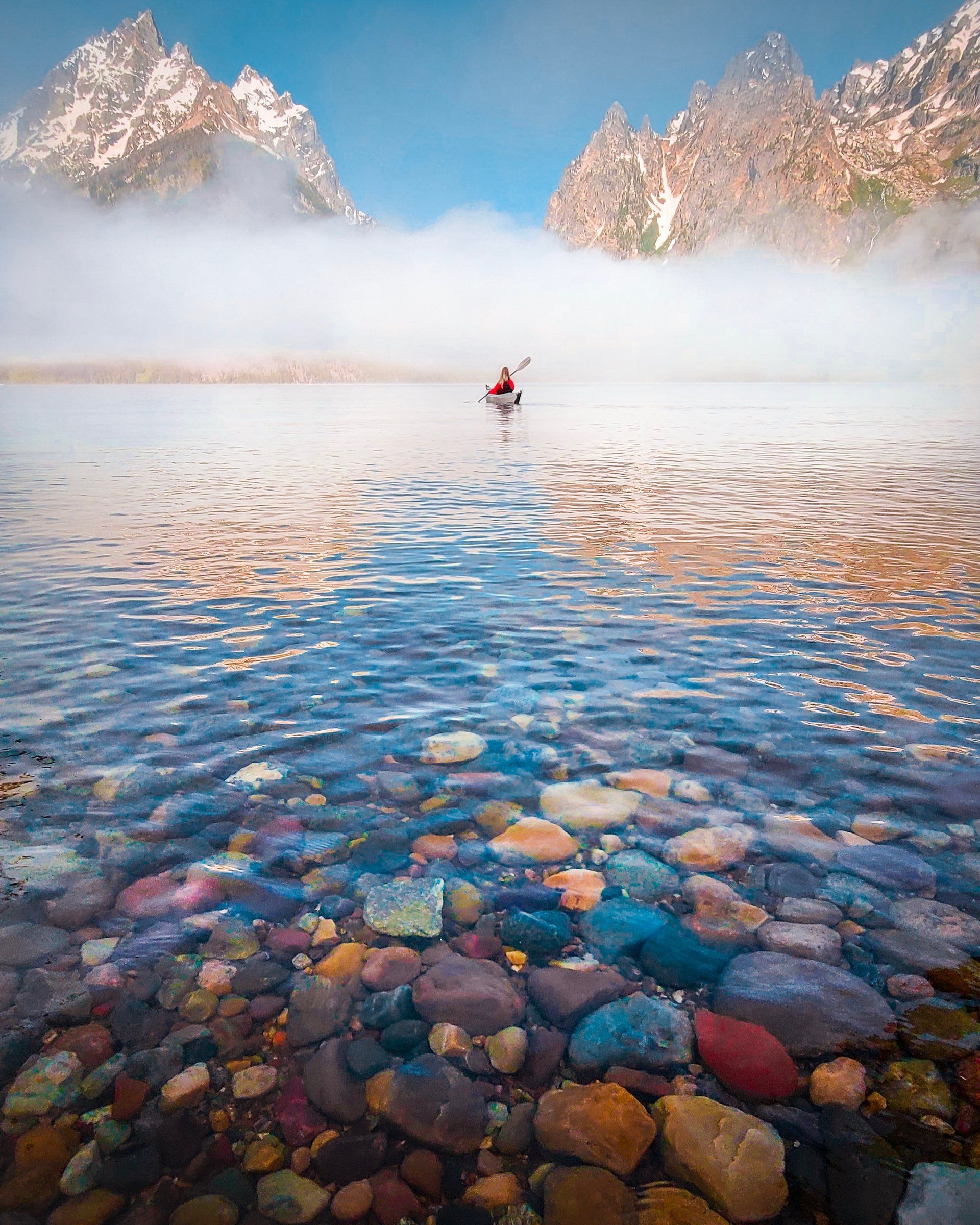 Kayaker paddling through morning fog on a clear lake over colorful stones, with jagged snowy peaks behind