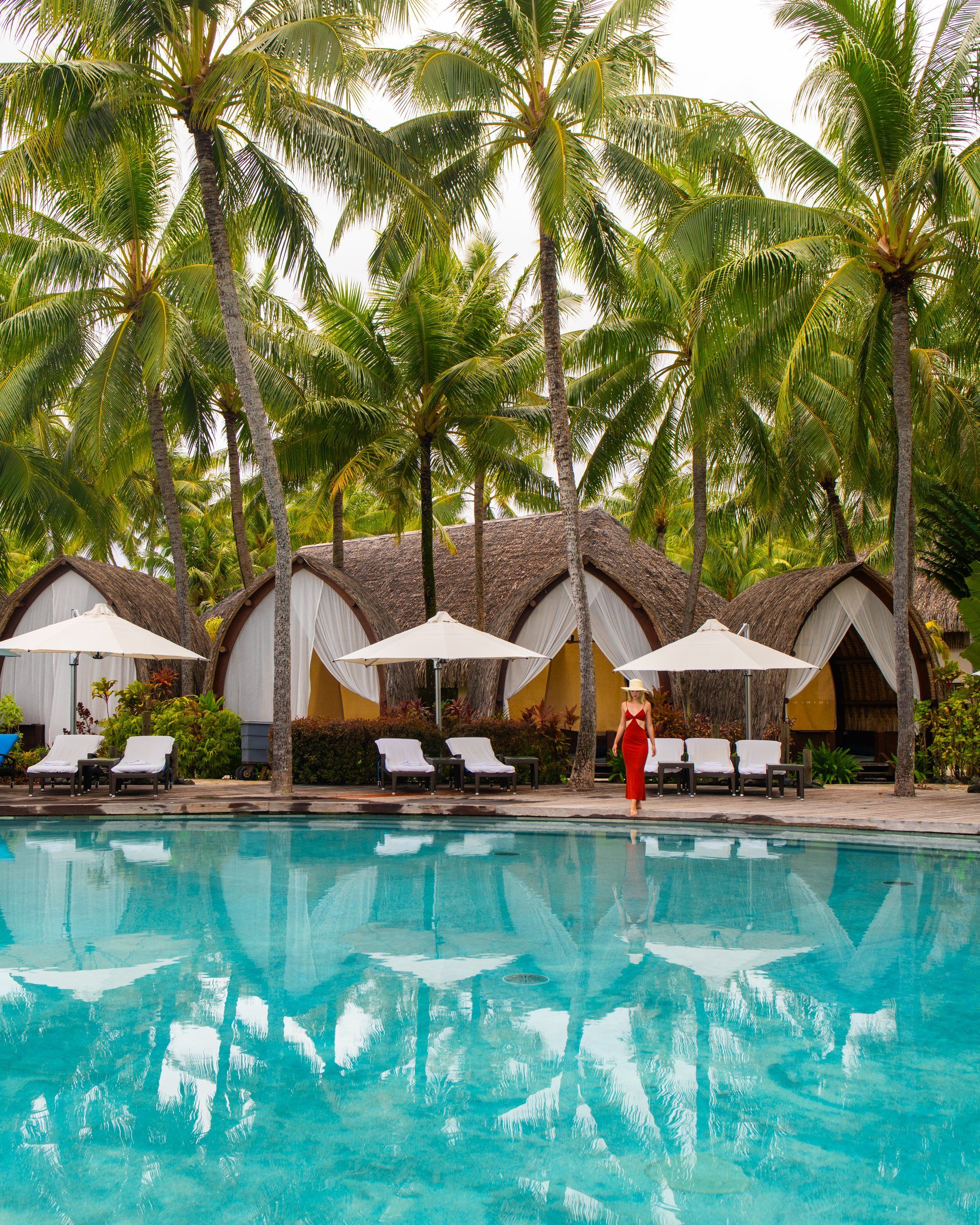 A woman in a red dress walking beside a palm-lined pool with thatched cabanas