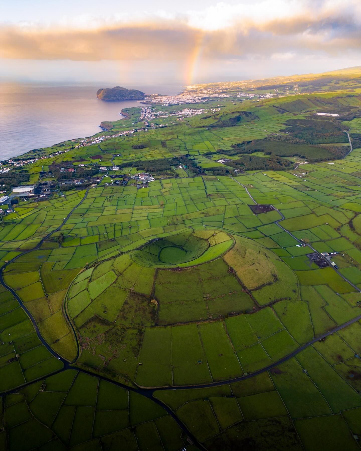 Wide aerial of Terceira's patchwork farmland around a crater, with a rainbow over the coast
