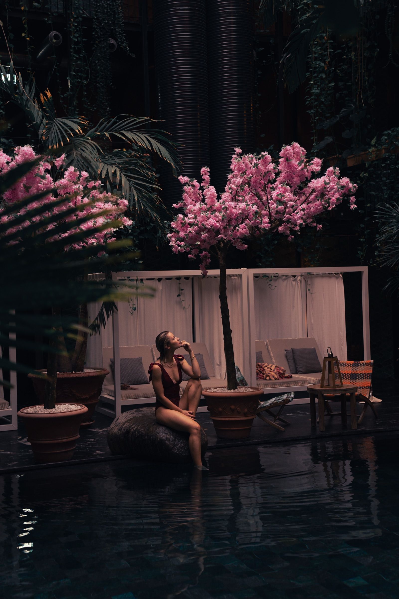 Woman seated by a dark pool under pink blossom trees, with cabanas behind