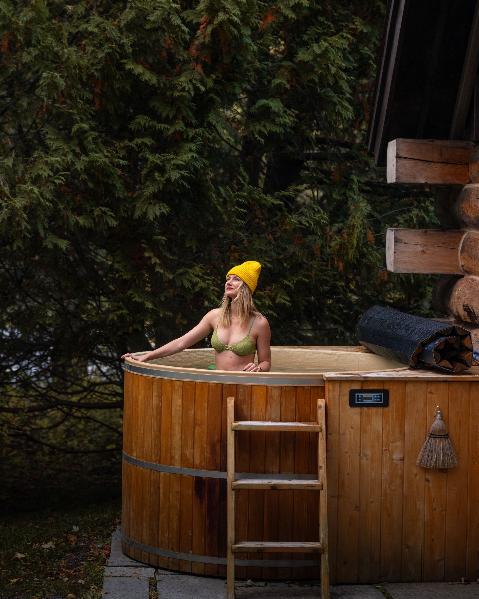 Woman in a yellow beanie in a wooden hot tub beside a log cabin