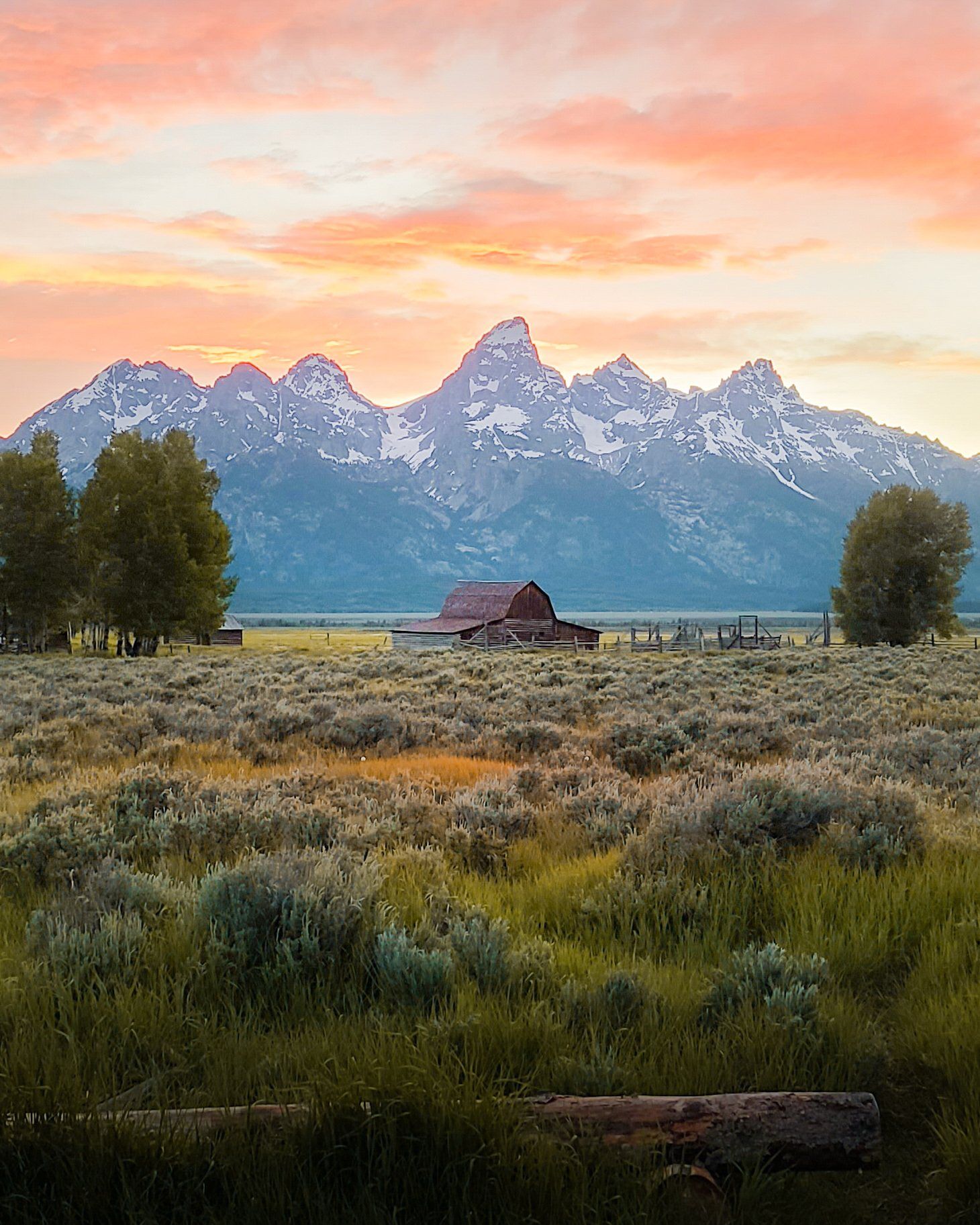 A weathered barn in sagebrush meadows below jagged peaks under a pink sunrise sky