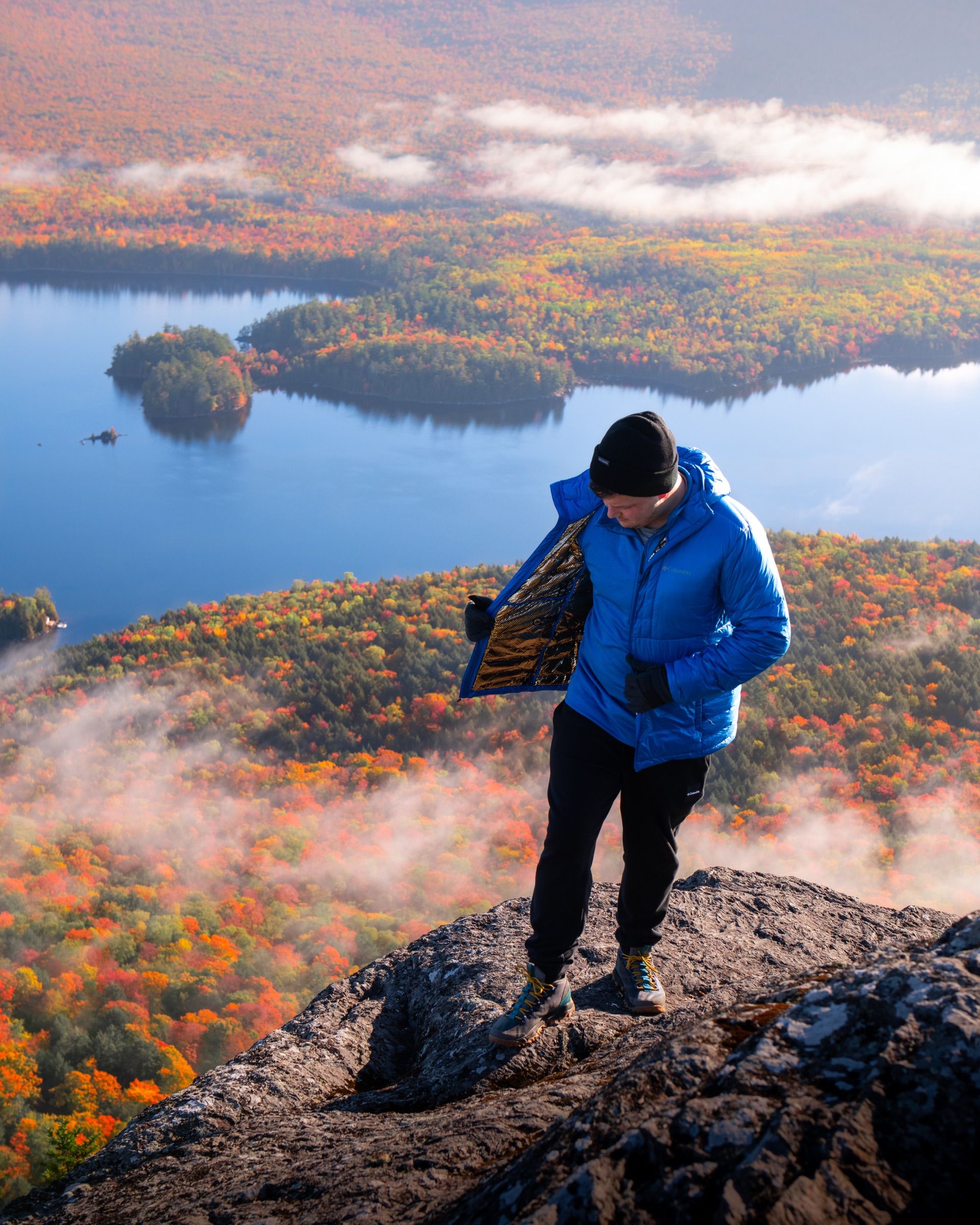 Man in a blue down jacket on a rocky summit above autumn forest, lakes and fog