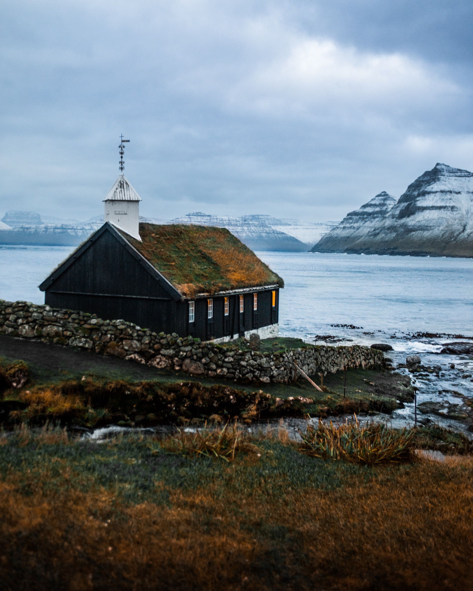 Black wooden church with a turf roof by a fjord, snowy mountains behind