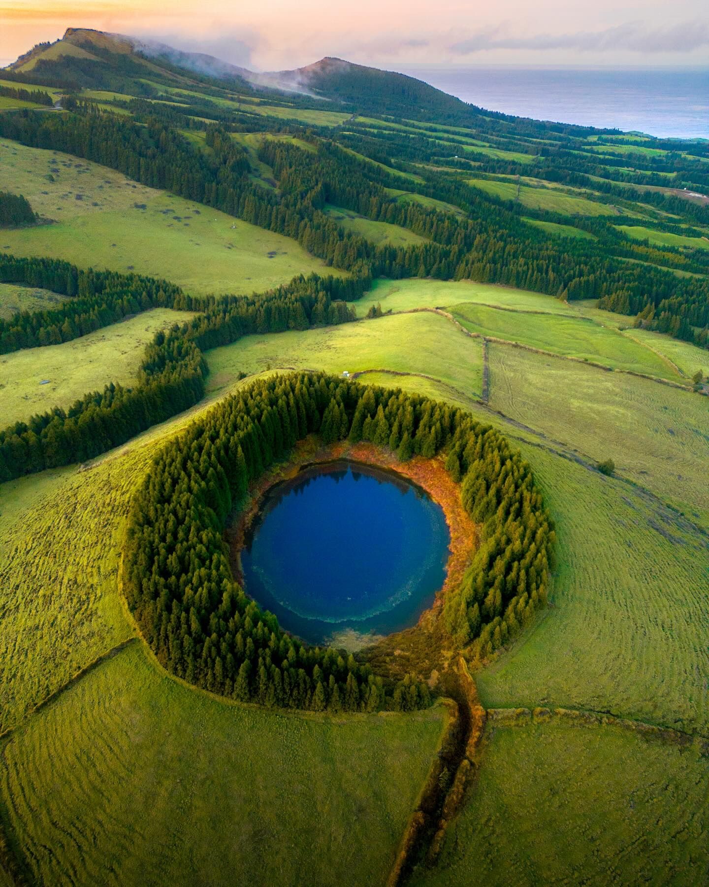 A deep blue crater lake ringed by forest in rolling green pasture