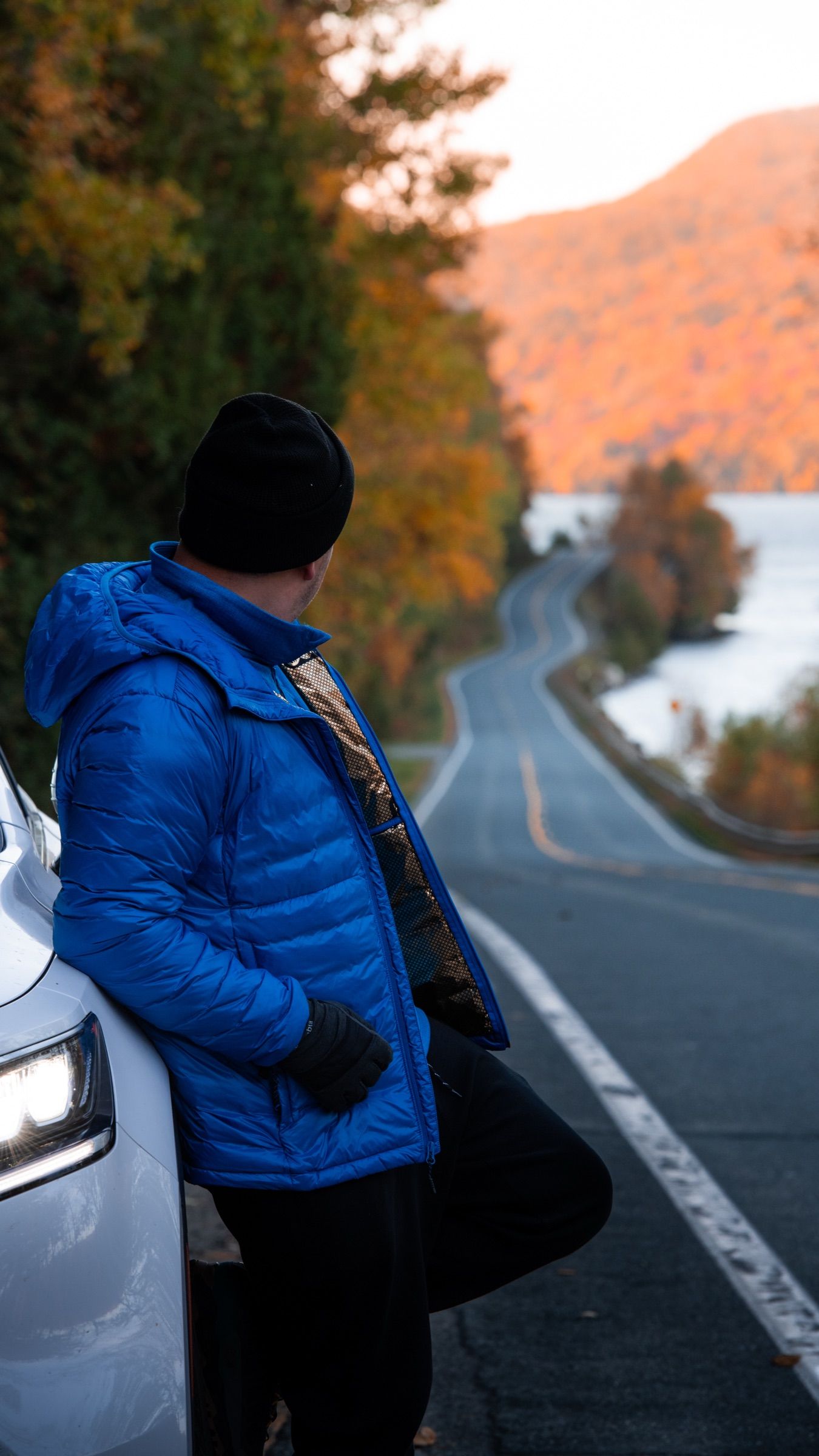 Man in a blue down jacket leaning on a car beside a winding lakeside road in autumn
