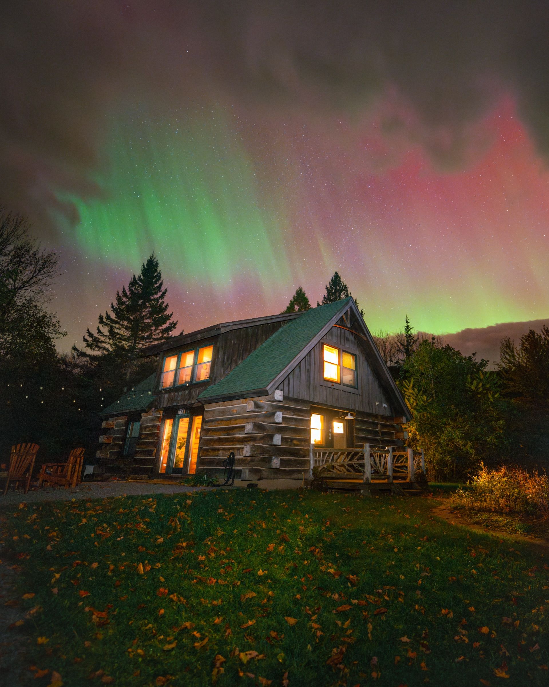 Log cabin with lit windows under green and pink northern lights