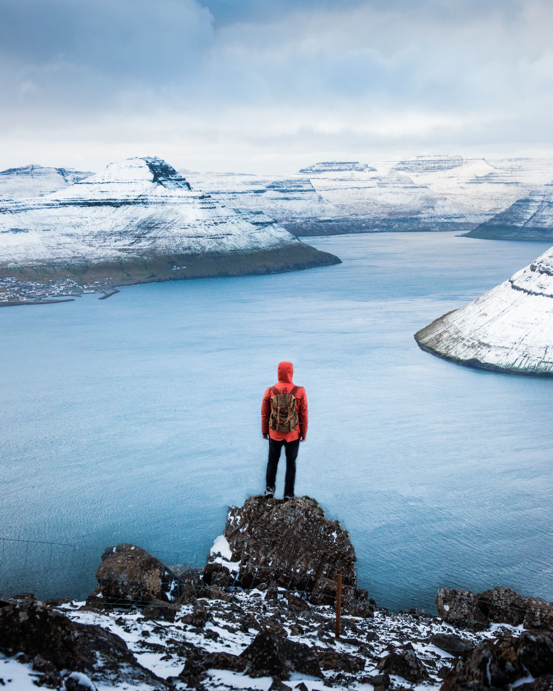 Hiker in a red jacket on a rock above a fjord with snow-covered mountains