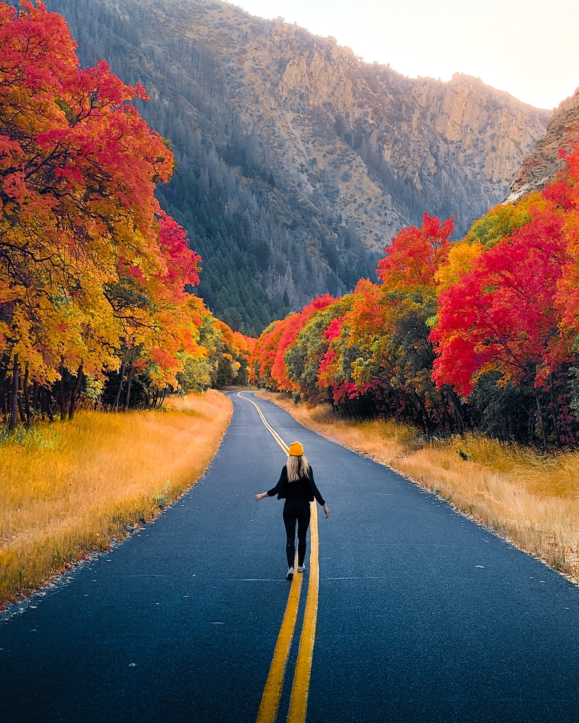 Woman walking down the center line of a canyon road lined with red and orange autumn trees