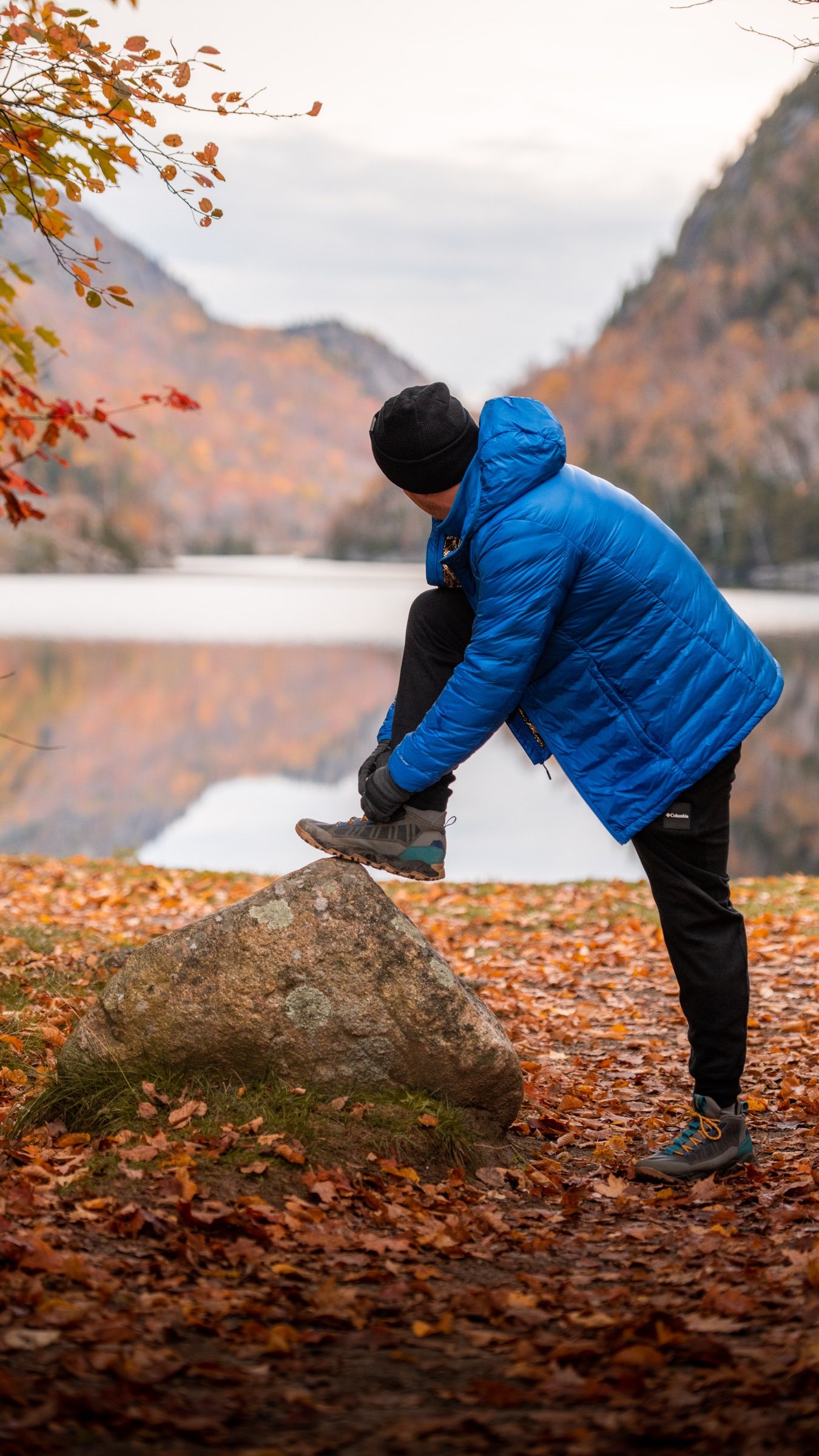 Man in a blue down jacket tying his boot on a rock by an autumn lake