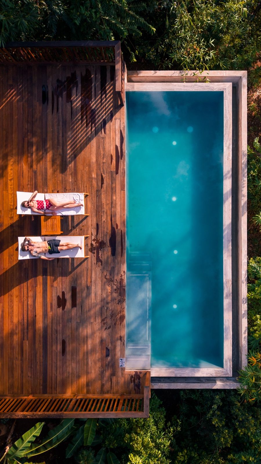 Top-down aerial of two people sunbathing beside a long plunge pool on a wooden deck