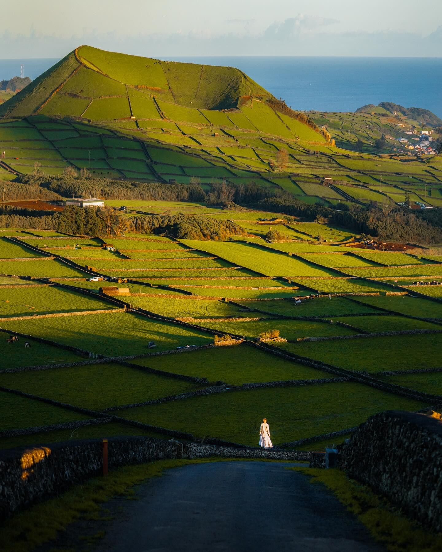A figure in white on a road below a grassy volcanic cone and stone-walled fields