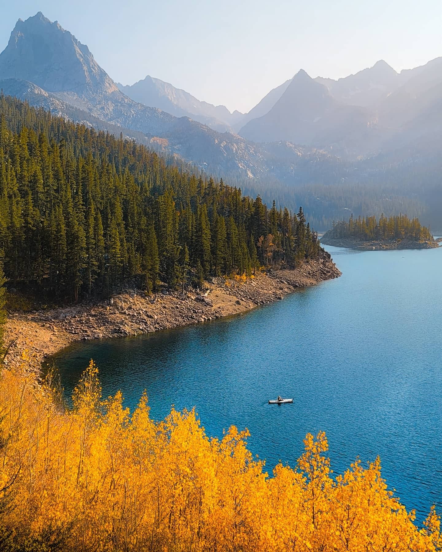 A paddleboarder on a blue alpine lake below hazy granite peaks, with golden aspens in the foreground