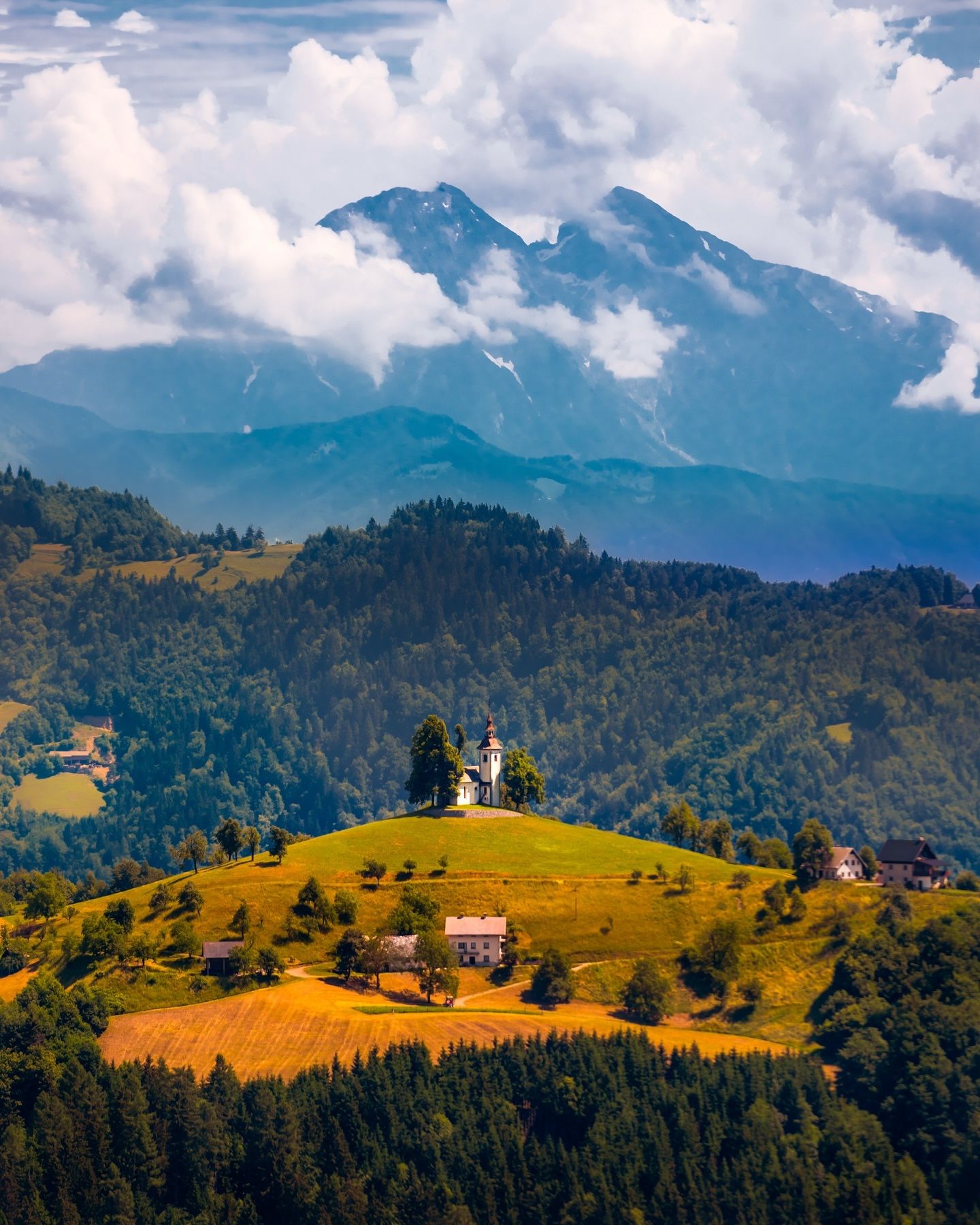 A white hilltop church above farmhouses and golden fields, with cloud-wrapped Alps behind
