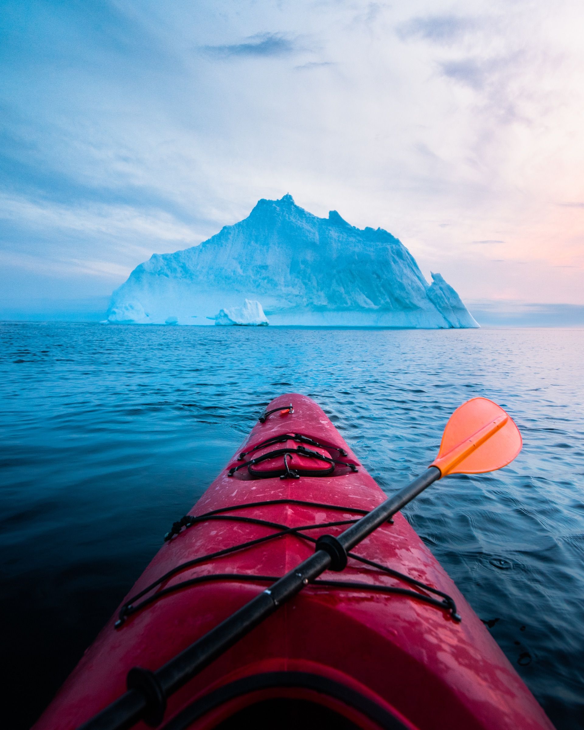 Bow of a red kayak and paddle facing a large iceberg