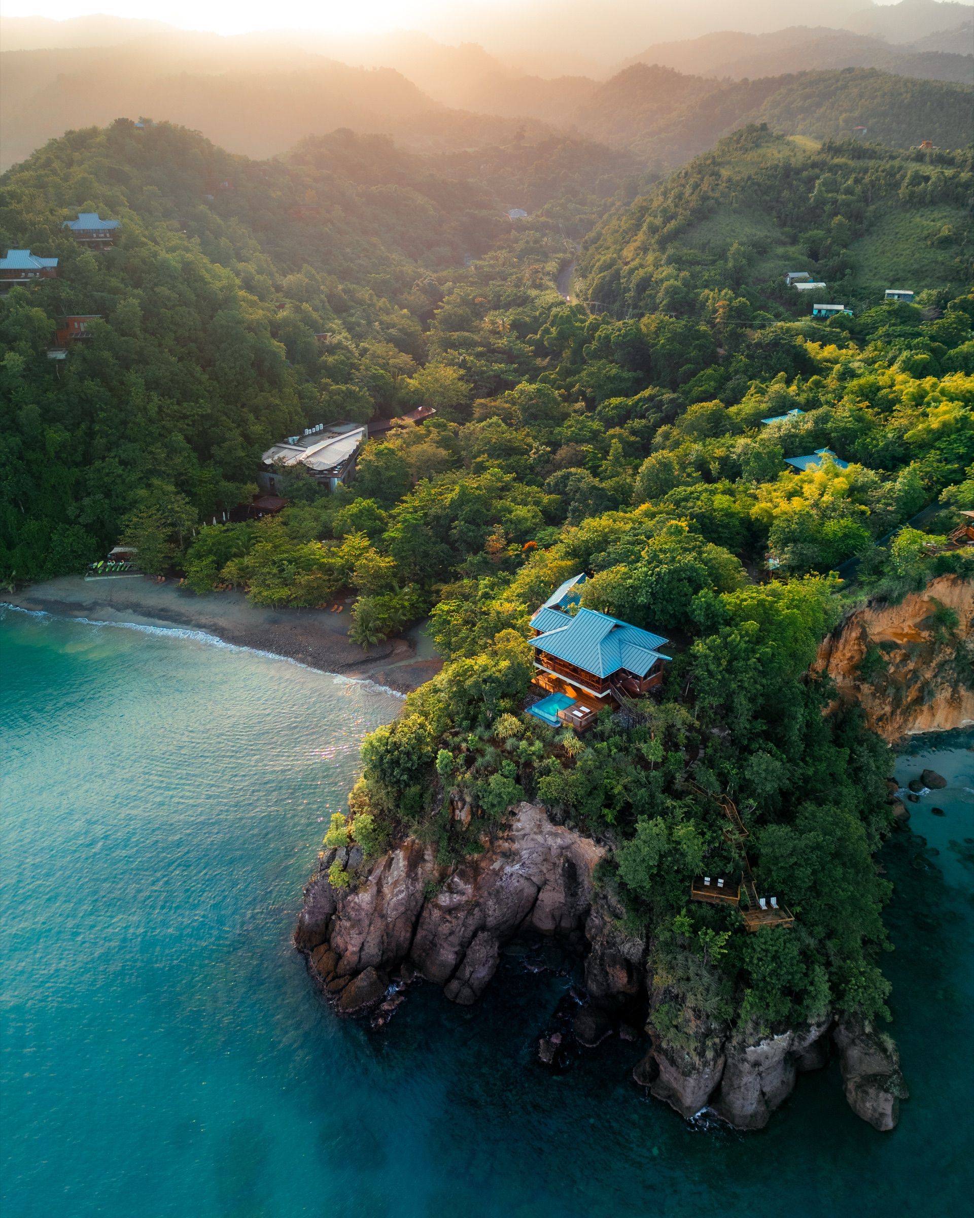 Aerial view of a villa on a rocky, forested point at sunrise, with green mountains behind