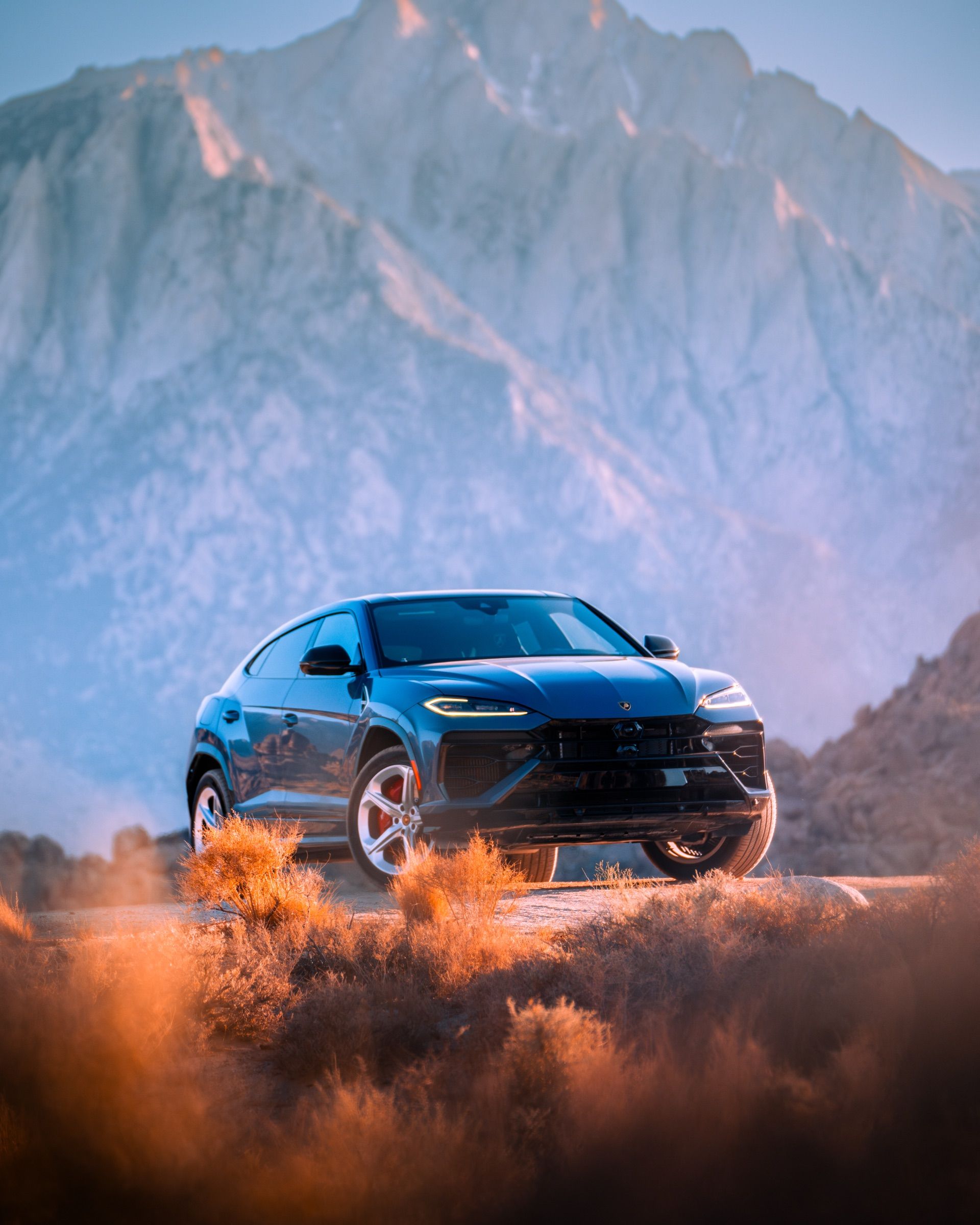 Blue Lamborghini Urus among desert brush below snowy granite peaks