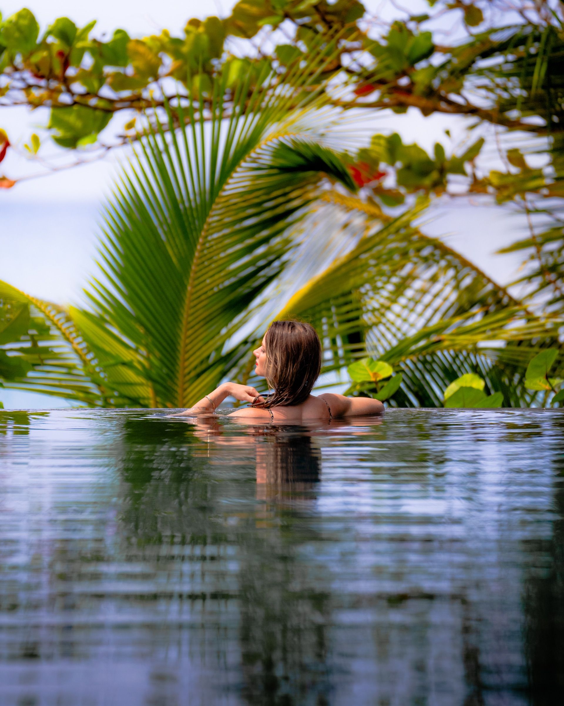 Woman in an infinity pool looking out past palm fronds