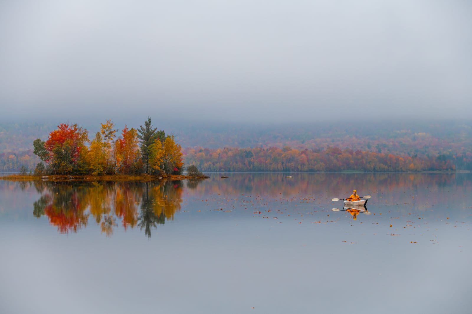 A small island of red and orange autumn trees reflected in a misty, glassy reservoir, with a lone canoe paddling nearby.