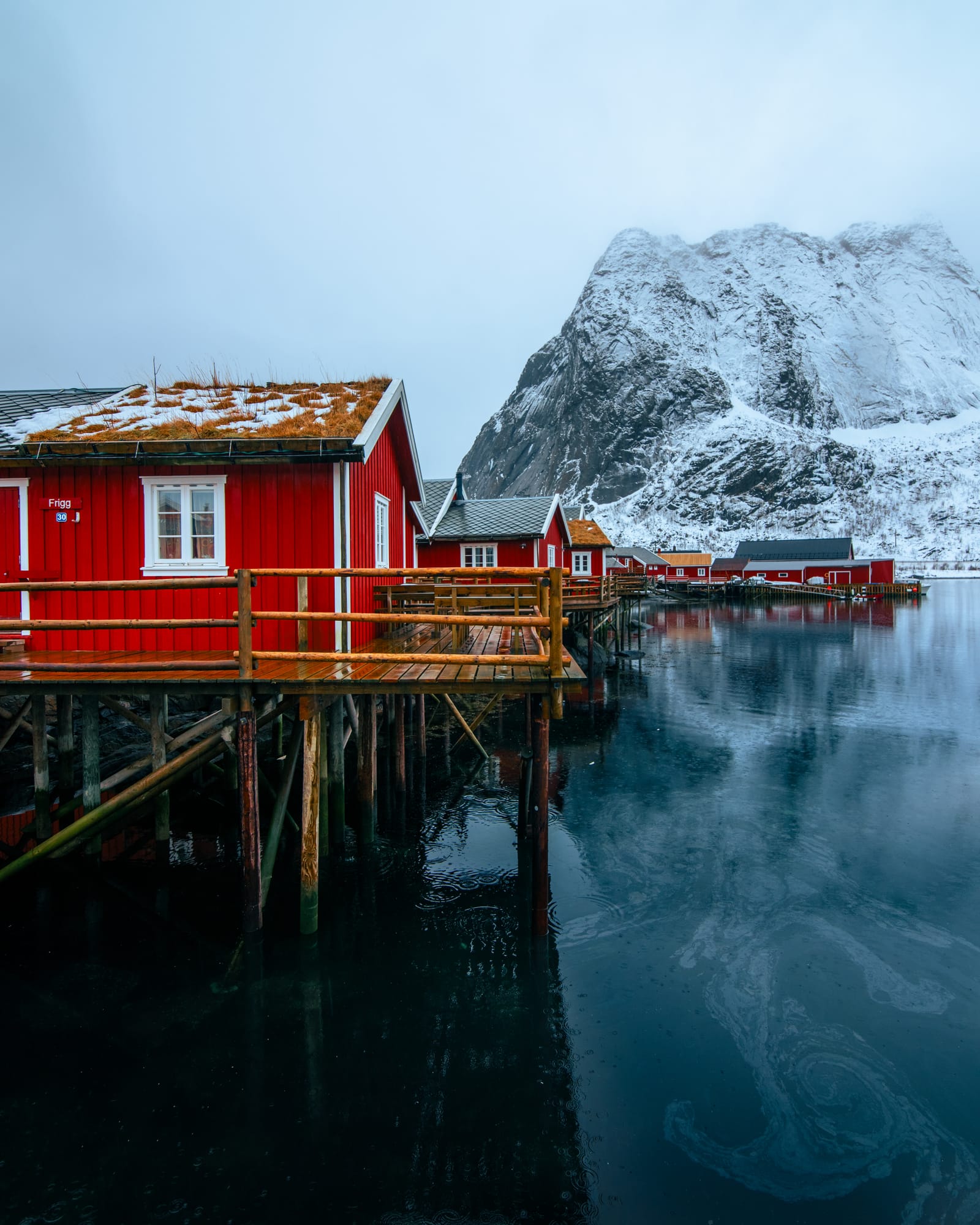 Red wooden fishing cabins on stilts over dark, still water beneath snowy mountains.